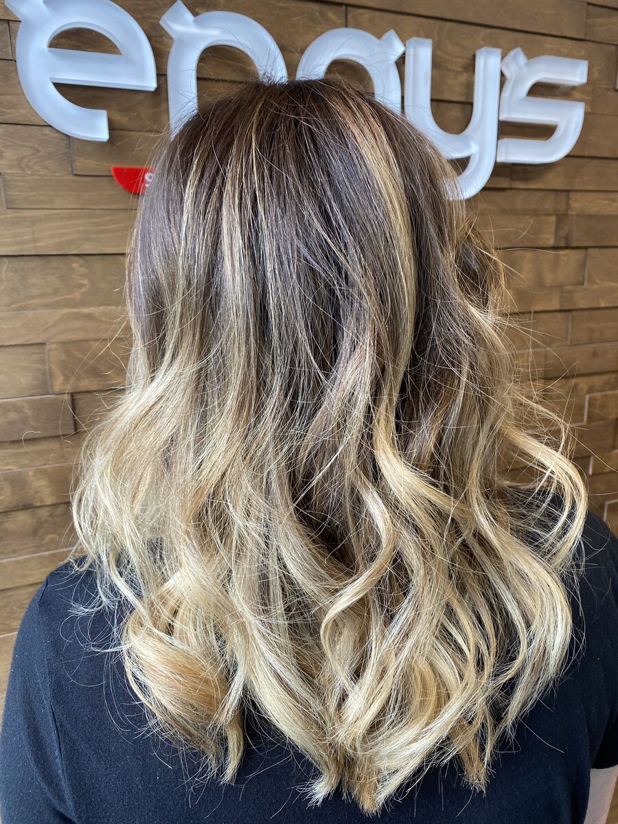 Blonde balayage on long waves, seen from the back in front of the Ergys Studio sign